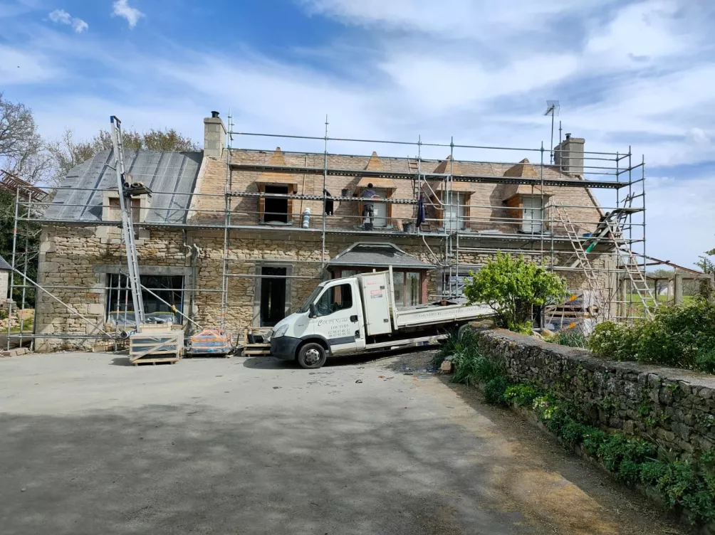 Chantier de toiture en cours sur une maison en pierre avec échafaudages et véhicule de chantier.